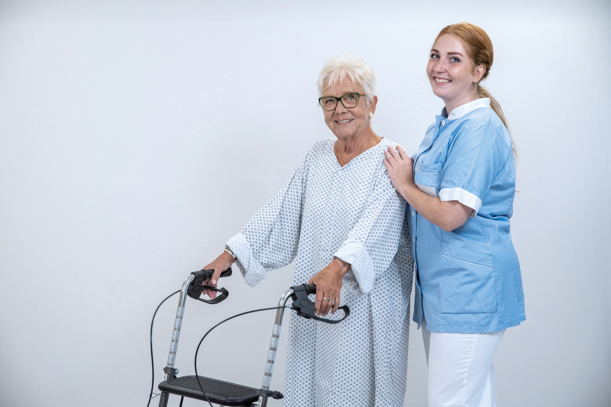 Eine ältere Dame mit Rollator im Krankenhausnachthemd, die von einer Gesundheits- und Krankenpflegerin unterstützt wird. 