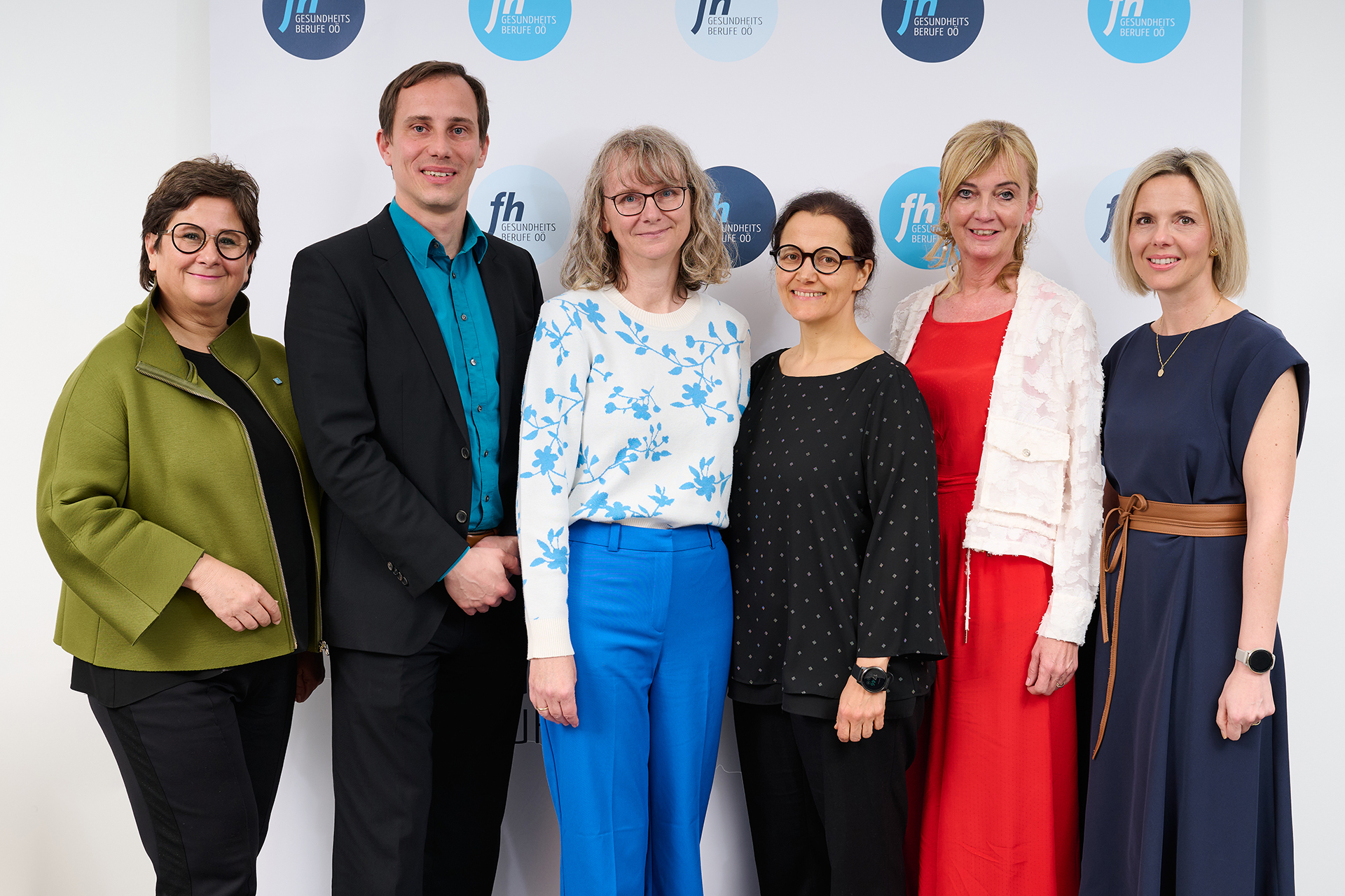 Sechs Personen stehen nebeneinander vor einer weißen Wand mit dem Logo der FH Gesundheitsberufe OÖ. Alle blicken in die Kamera. Von links: Eine Frau mit kurzem, dunklen Haar und Brille trägt eine olivgrüne Jacke über Schwarz. Ein Mann im schwarzen Anzug mit türkisfarbenem Hemd steht daneben. Die dritte Person, eine Frau mit schulterlangen hellen Haaren und Brille, trägt ein weißes Oberteil mit blauem Blumenmuster und eine blaue Hose. Neben ihr steht eine Frau mit dunkler Brille und schwarzem, gepunktetem Oberteil. Die fünfte Person trägt ein rotes Kleid und eine weiße, strukturierte Jacke. Ganz rechts steht eine Frau mit hellem Bob, dunkelblauem Kleid und braunem Gürtel 