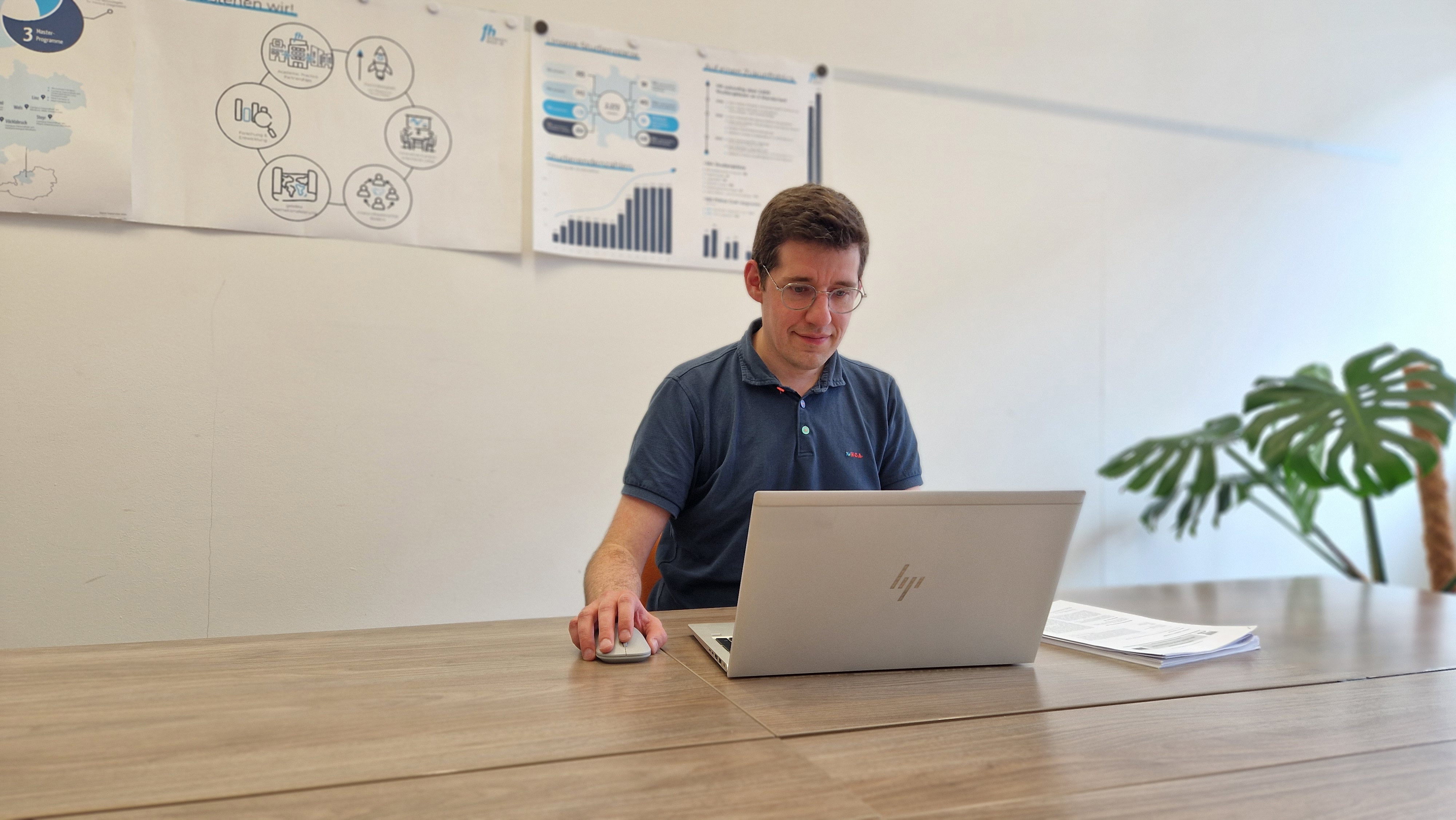 Ein Mann sitzt an einem Holztisch und arbeitet an einem silberfarbenen Laptop. Mit der rechten Hand bedient er eine Computermaus, die auf dem Tisch liegt. Er trägt ein dunkelblaues Poloshirt und eine Brille. Auf dem Tisch neben dem Laptop liegt ein Stapel Papiere. Im Hintergrund sind mehrere Poster mit grafischen Darstellungen und Diagrammen an der weißen Wand befestigt. Rechts im Bild steht eine grüne Zimmerpflanze mit großen Blättern. Fotocredit: FH Gesundheitsberufe OÖ 