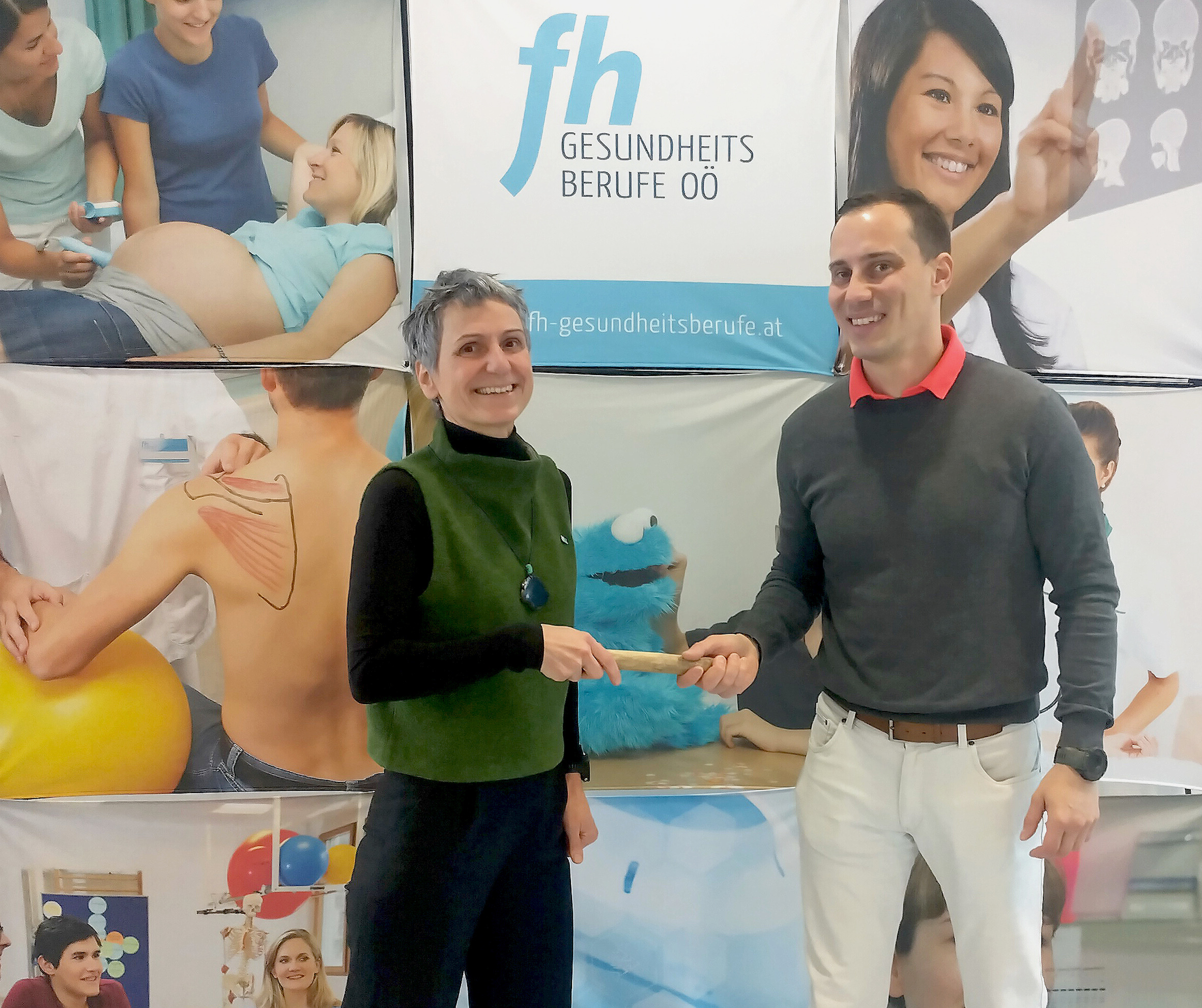Zwei Personen stehen vor einer Plakatwand mit Motiven der FH Gesundheitsberufe OÖ, links ist eine Frau mit hellem kurzem Haar, schwarzer Hose, schwarzem Oberteil und grünem Pullunder, sie überreicht dem Mann rechts neben ihr einen Staffelstab aus Holz. Der Mann hat dunkle kurze Haare. Über dem orangen Hemd trägt er einen grauen Pullover, dazu eine helle Hose  . 