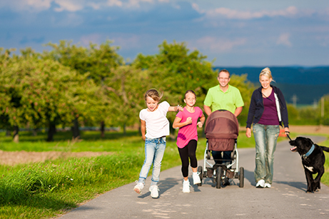 Eine Familie ist bei einem Spaziergang auf einem asphaltierten Weg in einer ländlichen Umgebung unterwegs. Zwei Kinder laufen lachend vorneweg. Eines trägt ein weißes T-Shirt und Jeans, das andere ein pinkes Shirt und eine schwarze Leggings. Dahinter gehen zwei Erwachsene: eine Person schiebt einen Kinderwagen, die andere führt einen großen schwarzen Hund an der Leine. Beide Erwachsenen lächeln. Im Hintergrund sind grüne Wiesen, Obstbäume und ein blauer Himmel mit wenigen Wolken zu sehen. Die Stimmung wirkt fröhlich und entspannt. 