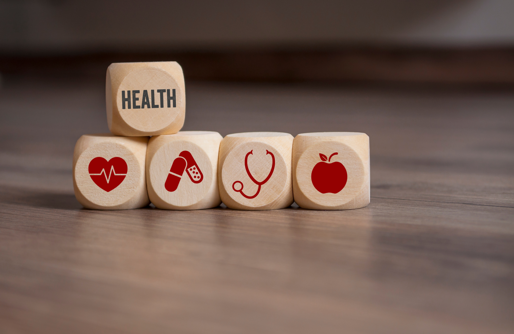 Fünf Holzwürfel, Vier liegen neben einander auf denen in roter Farbe jeweils ein Herz, Pillen, ein Stetoskop und ein Apfel abgebildet ist, ein Würfel liegt auf den anderen auf dem in schwarzer Schrift Health steht. 