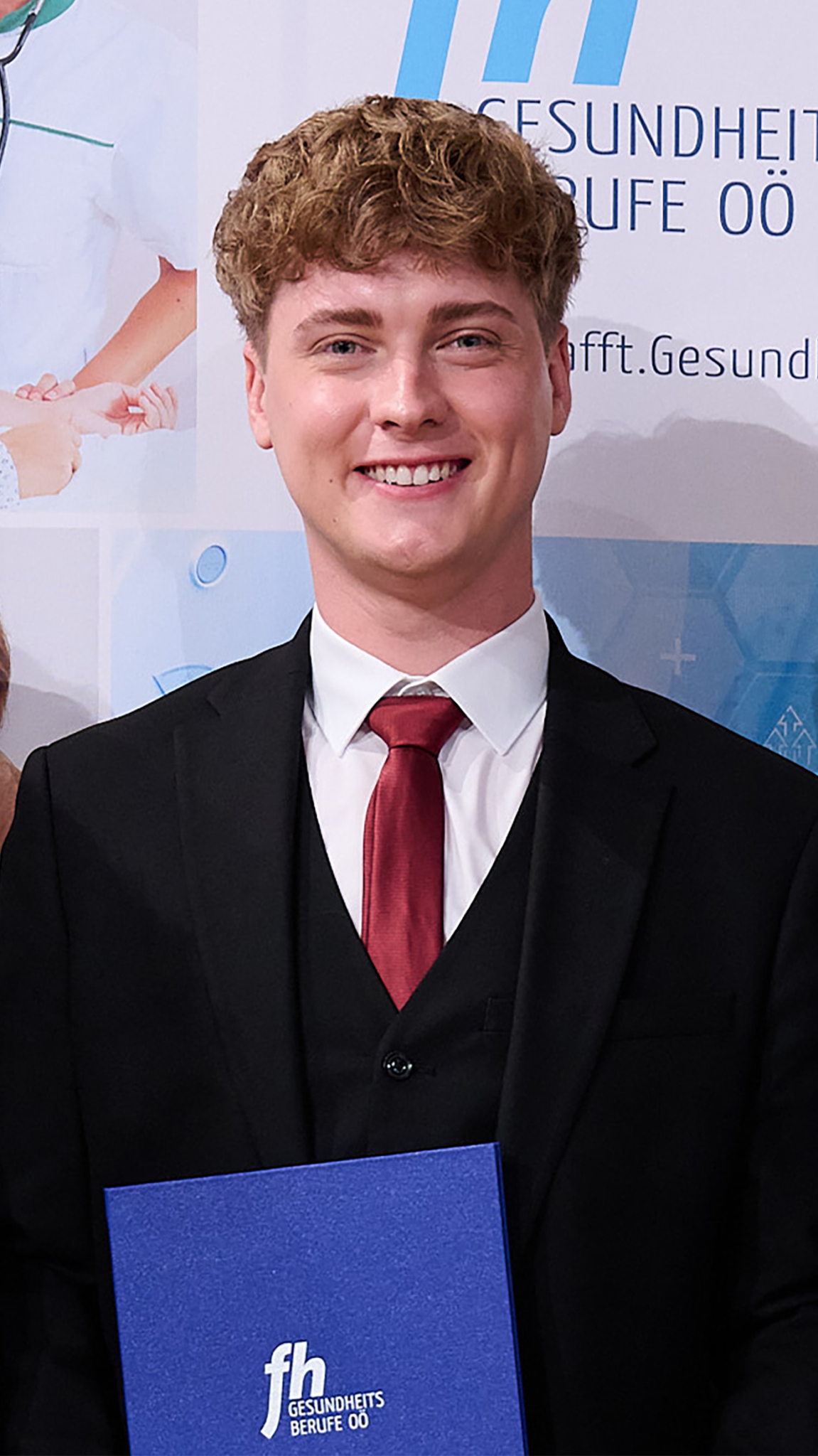 Junger Mann im Anzug mit Diplommappe der stolz in die Kamera lächelt 