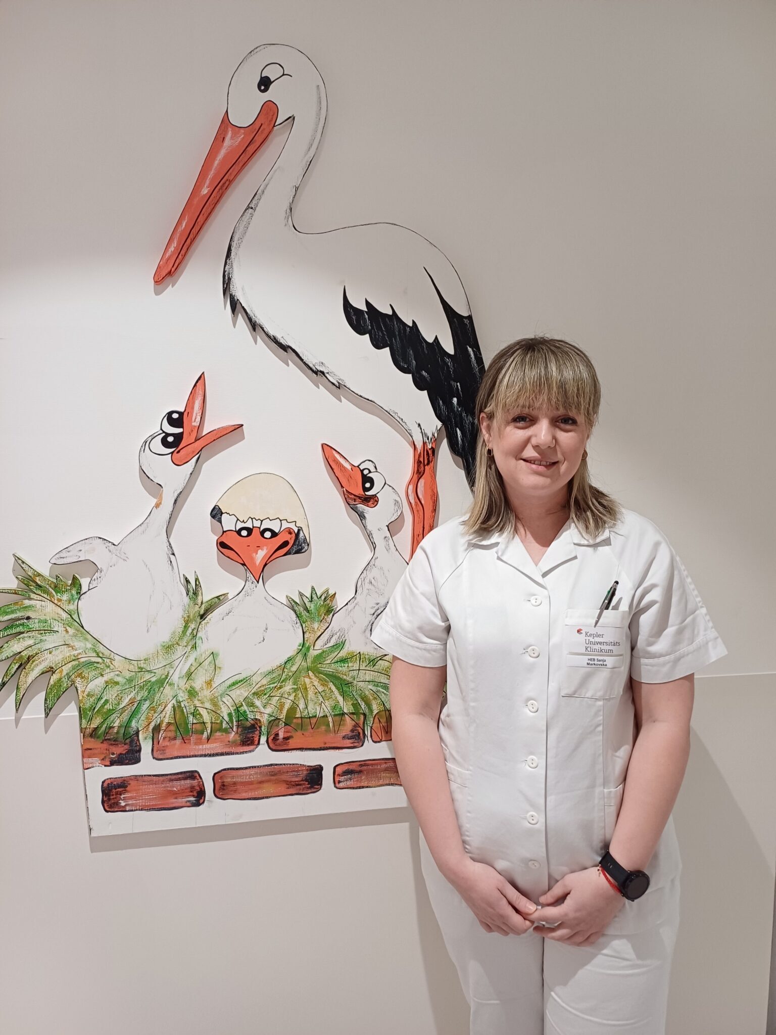 Eine Frau in weißer medizinischer Kleidung steht vor einer bunt bemalten Wand. Auf der Wand ist eine fröhliche Szene mit einem großen Storch und drei kleinen Störchen gemalt. Einer der kleinen Störche schlüpft gerade aus einem Ei. 